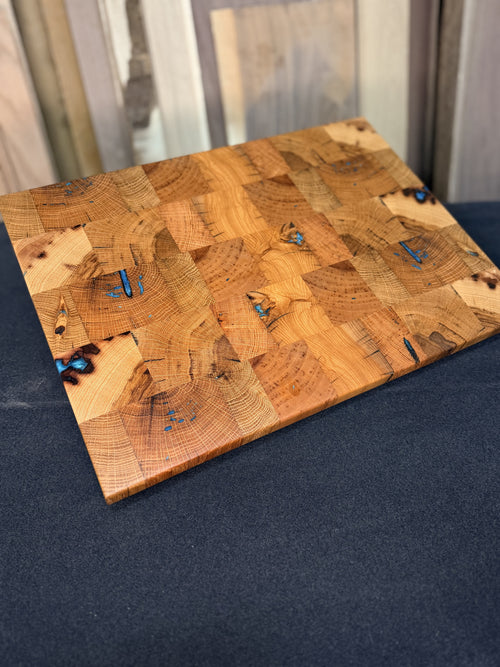 A rectangular wooden cutting board with a patchwork design of various natural wood slices and small blue inclusions, resting on a dark gray surface.