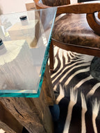 A close-up of a rustic wooden table with a clear glass top and a thick turquoise-edged glass layer, shown in a styled interior next to a brown leather chair and a black-and-white zebra print rug.