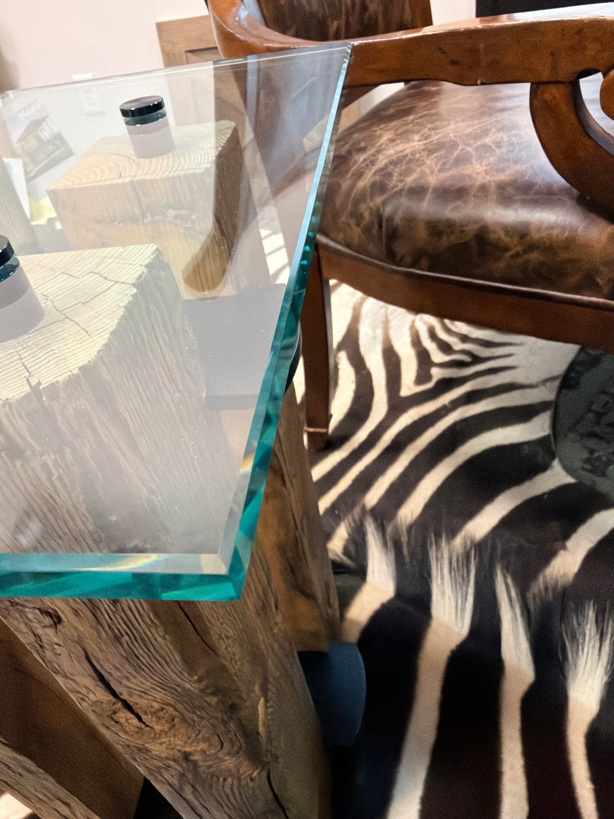 A close-up of a rustic wooden table with a clear glass top and a thick turquoise-edged glass layer, shown in a styled interior next to a brown leather chair and a black-and-white zebra print rug.